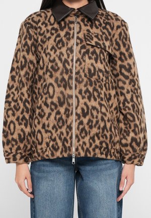 Mujer con una chaqueta marrón y negra con estampado de leopardo con cremallera, cuello de cuero negro y vaqueros azules.