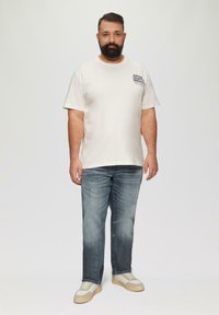 Beżowa bawełniana koszulka z graficznym nadrukiem, połączona z jasnoniebieskimi dżinsami. Buty akcentowane beżem i szarością, z teksturowaną powierzchnią.