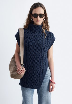 Femme portant un pull sans manches texturé bleu foncé, des lunettes de soleil, un jean bleu clair, des boucles d'oreilles anneaux en argent, des bagues, et portant un sac à bandoulière en daim beige.