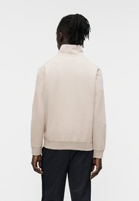 Μπεζ φούτερ με ψηλό γιακά, ριμπ μανσέτες και λεία υφή. Φοριέται με σκούρο παντελόνι, σε θέα από πίσω.