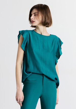 Una donna in una blusa senza maniche teal con volant e pantaloni abbinati si trova davanti a uno sfondo semplice, guardando verso sinistra.