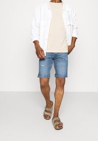 Pantalones cortos de mezclilla en azul claro con bordes deshilachados, combinados con una camiseta beige y una camisa blanca de botones. Sandalias grises completan el conjunto.