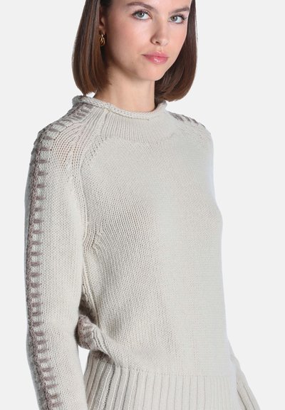 Femme aux cheveux bruns courts portant un pull en tricot crème avec des détails de bandes brunes texturées le long des manches et des épaules.