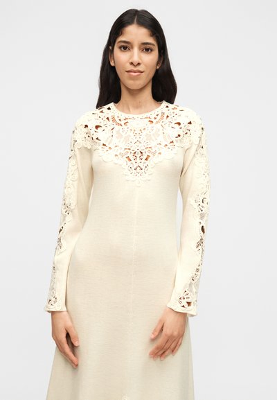 Γυναίκα που φοράει εκρού φόρεμα με μακριά μανίκια και περίτεχνη δαντέλα στο στήθος και στα μανίκια, στέκεται μπροστά σε απλό ανοιχτόφωτο φόντο.