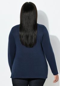 Laivastonsininen neulepaita, jossa on pitkät hihat ja rento istuvuus, teksturoidulla kankaalla ja pyöristetty helmalla, yhdistetty tummiin farkkuihin.