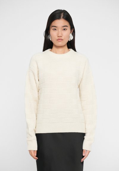 Pull en tricot beige avec un motif texturé, à manches longues et col rond, associé à une jupe noire.