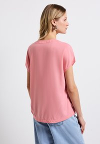 Krátký rukáv, růžová blůza z hladké, lehké tkaniny. Má kulatý výstřih a volný střih, stylizovaný s lehkými džíny.