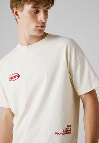 Cremefarvet bomulds t-shirt med korte ærmer og rund hals. Røde broderede logoer på venstre bryst og nederste side. Glat tekstur.