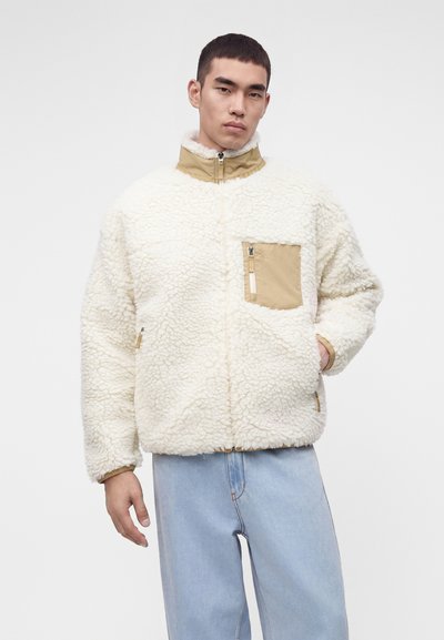 Νέος άνδρας που φοράει λευκό σακάκι sherpa fleece με τσέπη και γιακά σε ταμπ χρώμα, συνδυασμένο με ανοιχτό μπλε τζιν, στέκεται μπροστά σε απλό φόντο.