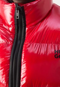 Veste matelassée rouge brillante avec col haut et fermeture éclair noire, affichant partiellement le logo "HUGO" sur la poitrine.