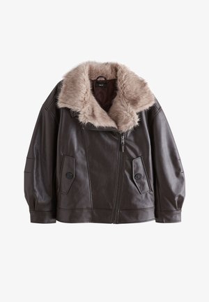 Donna che indossa una giacca di pelle marrone scuro con un grande colletto in pelliccia sintetica beige chiaro, mentre sistema la cerniera frontale, su sfondo grigio.