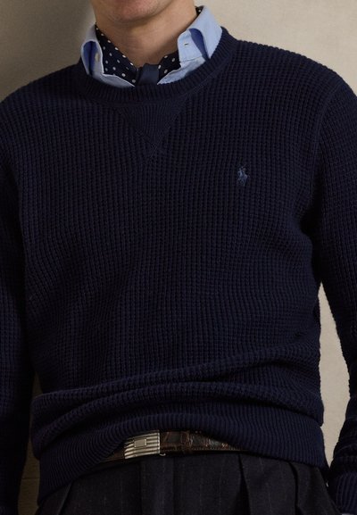 Μπλε ναυτικό πλεκτό πουλόβερ με ραβδωτή υφή και σχέδιο με λαιμόκοψη V. Διαθέτει μικρό λογότυπο στο στήθος και συνδυάζεται με ανοιχτό μπλε πουκάμισο με γιακά.