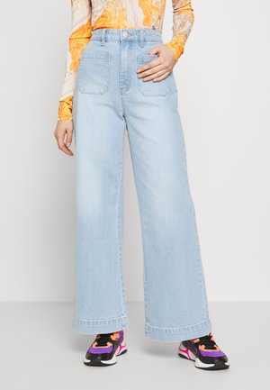 Persona con vaqueros de pierna ancha de color azul claro, camiseta de manga larga naranja con estampado y zapatillas negras, rosas y moradas.