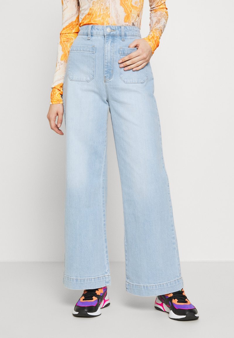 Personne portant un jean large bleu clair, un haut à manches longues orange à motifs, et des baskets noires, roses et violettes.