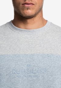Sweat-shirt gris avec une bande horizontale bleu clair, texture douce, col côtelé et texte du logo brodé sur la poitrine.
