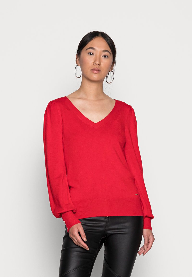 Roter Strickpullover mit V-Ausschnitt und langen, gerafften Ärmeln. Mit geripptem Saum und leichtem Glanz. Kombiniert mit schwarzen Lederhosen.