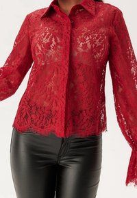 Blouse en dentelle rouge avec col, manches longues et texture transparente. Dotée de motifs floraux complexes et d'un ourlet droit. Associée à un pantalon noir.
