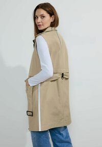 Beige ärmelloser Trenchcoat mit hohem Kragen, Seitentaschen und einem schwarzen Schnallendetail auf der Rückseite. Weiße Streifen entlang der Seiten.