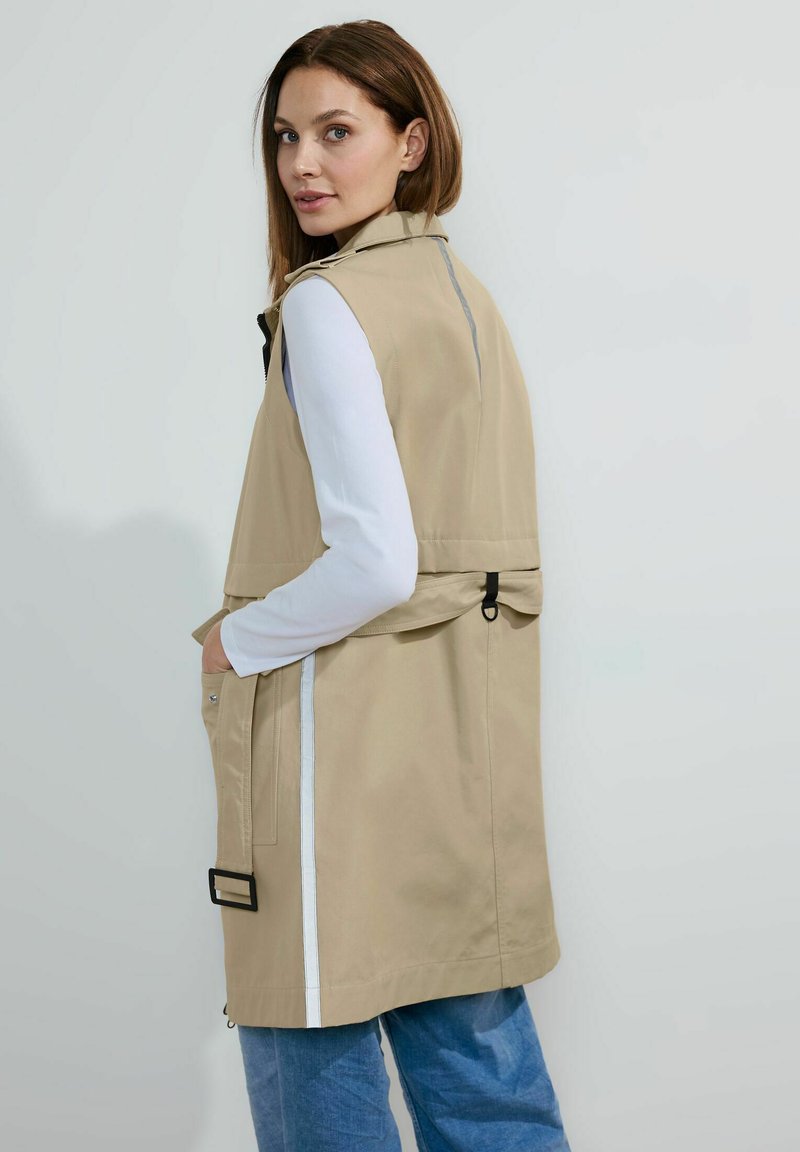 Beige ärmelloser Trenchcoat mit hohem Kragen, Seitentaschen und einem schwarzen Schnallendetail auf der Rückseite. Weiße Streifen entlang der Seiten.