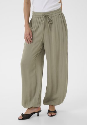 Pantalon ample couleur olive clair avec taille élastique et cordon de serrage, porté avec des sandales à talons noires ouvertes et un haut blanc.