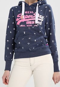 Felpa navy con logo "Superdry" rosa, cordini bianchi e piccoli motivi bianchi. Presenta una tasca frontale e polsini a costine. Materiale morbido.