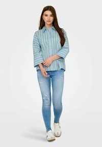 Camicia azzurro chiaro con righe verticali, vestibilità comoda e maniche ampie, abbinata a jeans azzurro chiaro strappati e sneaker bianche.