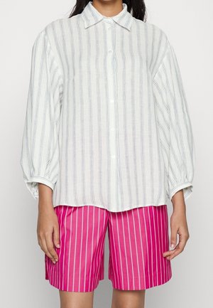 Žena má na sobě volnou bílou košili s černými svislými pruhy a jasně růžové šortky po kolena s bílými svislými pruhy.