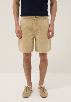 Uomo con pantaloncini beige al ginocchio, scarpe beige stringate e camicia nera in piedi davanti a uno sfondo uniforme.
