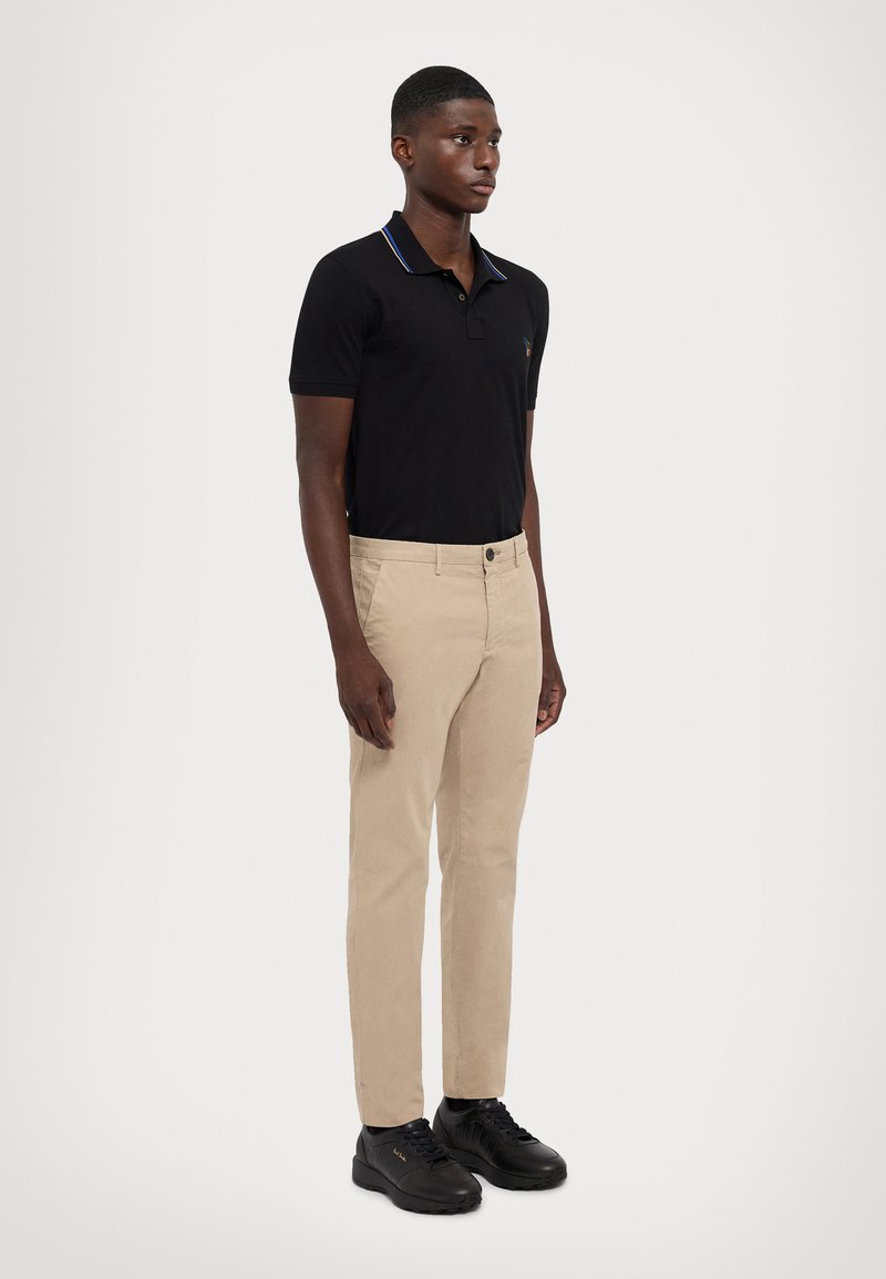 Camisa polo preta com colarinho em contraste, calças chino bege e ténis pretos. Ajuste casual; textura ribbed no colarinho e tecido suave nas calças.