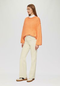 Maglione over-size arancione, jeans a zampa di elefante crema e scarpe beige con fasce nere; trame morbide, vestibilità rilassata e look a strati.