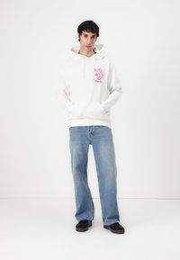 Witte hoodie met roze grafische afbeelding op de borst, gecombineerd met lichtblauwe losvallende jeans en zwarte schoenen.