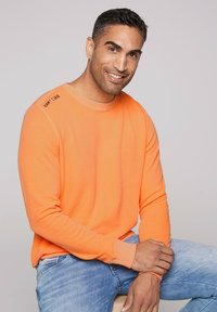Oranje blouse met lange mouwen van zachte stof, met een ronde halslijn en een logo op de linkerschouder. In combinatie met blauwe jeans.