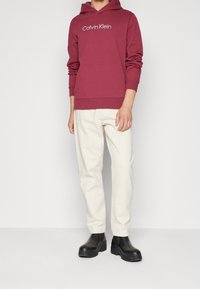Burgundowa bluza z kapturem z logo "Calvin Klein", białe kremowe spodnie i matowe czarne botki do kostki z grubymi podeszwami. Prosty, casualowy design.