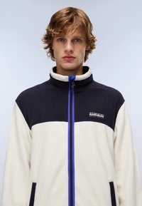 Giovane uomo con capelli castano chiaro mossi, che indossa una giacca in pile bianca e blu marino con una cerniera blu e un logo sul petto.
