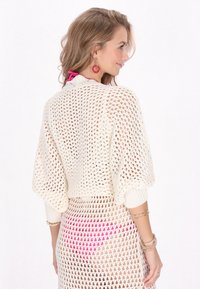 Cardigan bianco in maglia aperta con maniche larghe, che mostra una trama adatta. La modella indossa un accento rosa sotto, abbinato a gioielli dorati.