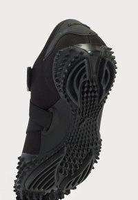 Zapato deportivo negro con suela de goma texturizada, parte superior de malla y correa de Velcro. Presenta púas de tracción multidireccional para una mayor adherencia.