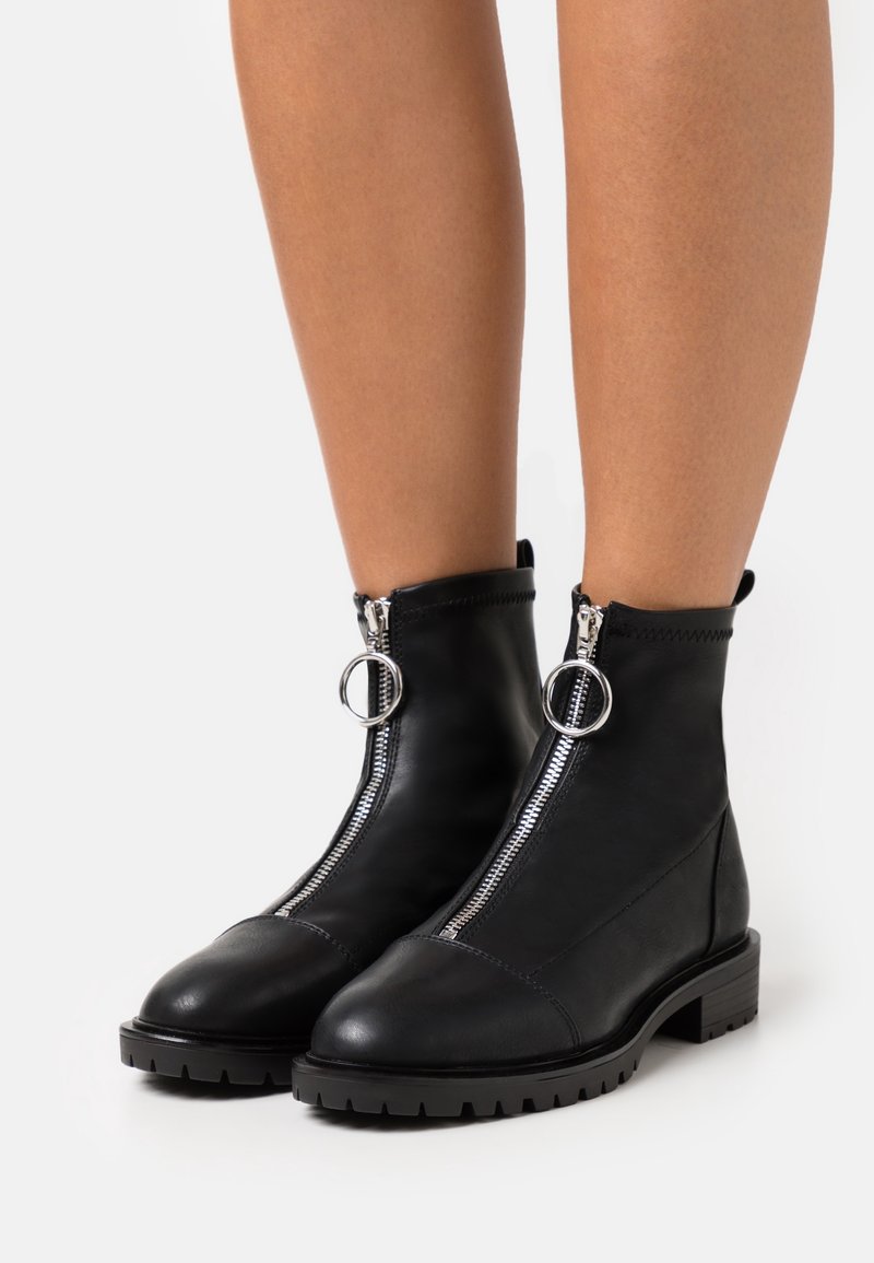 Bottines noires à cheville avec fermeture éclair argentée à l'avant et anneaux ronds, portées sur des jambes nues, debout sur fond blanc.