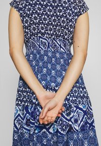 Femme aux mains croisées dans le dos, portant une robe bleue à motifs géométriques et floraux sur un fond clair.