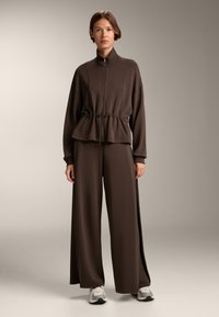 Giacca marrone con zip e cintura regolabile in vita, abbinata a pantaloni larghi coordinati, realizzata in un tessuto morbido, indossata con sneakers chiare.