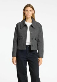 Giacca crop grigia a spina di pesce con colletto, bottoni frontali e due tasche laterali, abbinata a jeans neri a vita alta.