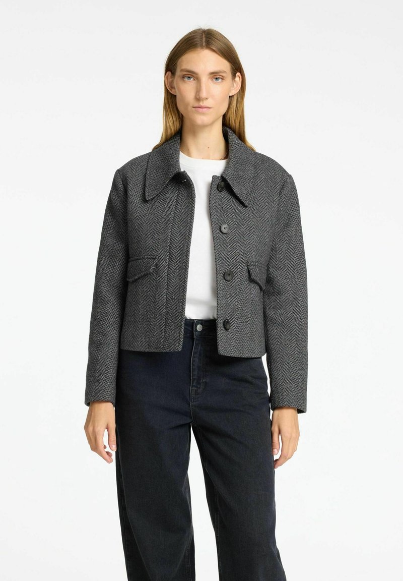 Giacca crop grigia a spina di pesce con colletto, bottoni frontali e due tasche laterali, abbinata a jeans neri a vita alta.