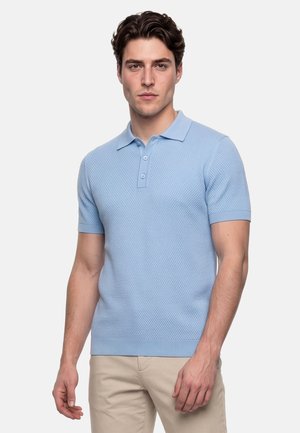 Jonge man draagt een lichtblauw gestructureerd gebreid poloshirt en beige broek, staand tegen een effen lichte achtergrond.