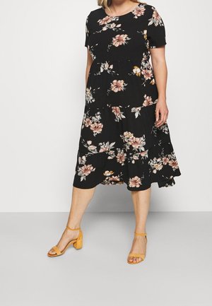 Kvinde iført en sort midi-kjole med pink og hvide blomsterprint og sennepsgule blokhælssandaler, stående mod en ensfarvet baggrund.