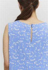 Blouse bleue à motifs floraux avec une ouverture en forme de goutte au dos, présentant des motifs blancs subtils et une fermeture à bouton au niveau du col.