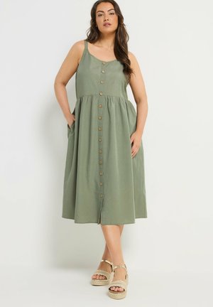 Femme portant une robe midi boutonnée vert olive à fines bretelles et des sandales plateformes beiges, debout devant un fond blanc.