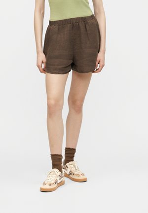 Sötétbarna elasztikus derékpántú rövidnadrágot viselő személy, barna zoknival és állatmintás részletekkel díszített bézs tornacipőben, fehér háttér előtt.