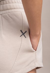 Beige katoenen shorts met een gestructureerde afwerking, voorzien van een zijzak en een donkergrijze geborduurde "X" accent op de rand van de zak.