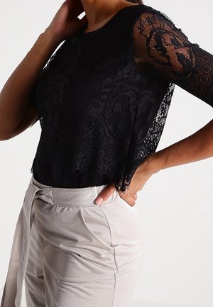 Kvinde iført en sort blonde top med korte ærmer, stoppet ind i højtaljede beige bukser med en bundet bælte, poserende med den ene hånd på hoften.