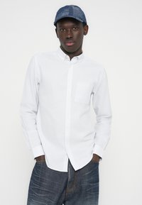 Νεαρός άντρας που φοράει μπλε καπέλο με σχέδια, λευκό πουκάμισο με ρίγες και φαρδιά σκούρα μπλε τζιν, στέκεται με τα χέρια στις τσέπες.