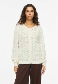 Femme aux longs cheveux bouclés portant un cardigan en maille crème avec des boutons en perle et des bords festonnés, associé à un pantalon large marron foncé.